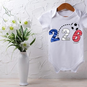 Peut inclure: Body bébé blanc avec un ballon de foot et le chiffre "226" aux couleurs du drapeau français. Le mot "France" est sous le chiffre 6. Le body est suspendu à un cintre en bois.