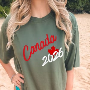 May include: Olive green t-shirt with the word "Canada" in red script, a red maple leaf, and the number "2026" in white. The shirt is a casual, short-sleeved style.