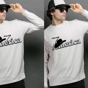 Peut inclure: Sweat-shirt blanc à manches longues avec un motif noir d'un chapeau de cowboy et le mot "America". Le sweat-shirt est porté avec une casquette de camionneur noire et blanche et des lunettes de soleil.