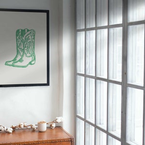 May include: A framed print featuring a green outline of a pair of cowboy boots. The artwork is set against a light background and framed in black. The print hangs on a white wall, with a window to the right.