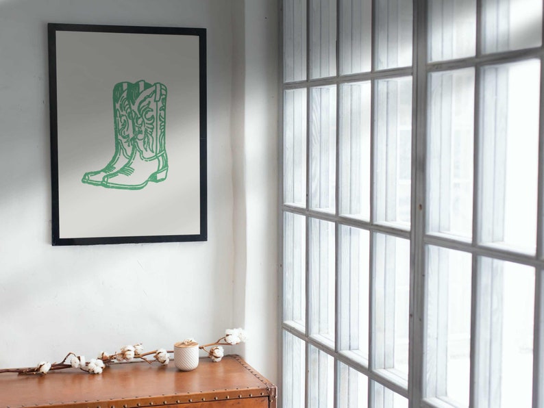 May include: A framed print featuring a green outline of a pair of cowboy boots. The artwork is set against a light background and framed in black. The print hangs on a white wall, with a window to the right.
