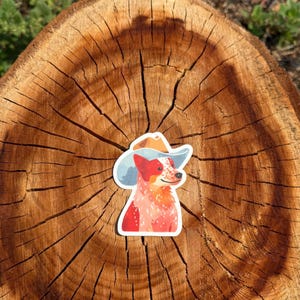 May include: A die-cut sticker featuring a red and white speckled dog wearing a light blue cowboy hat. The dog has a pointed snout, dark eyes, and a small black nose, rendered in a flat, illustrated style with a white border around the edges.