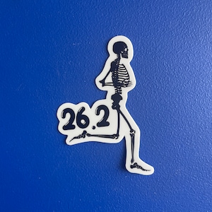 May include: A die-cut sticker featuring a black skeleton in a running pose with the text 26.2 in a bold, rounded font. The skeleton is depicted in profile, showing the rib cage, spine, and leg bones, with a white border outlining the entire design.
