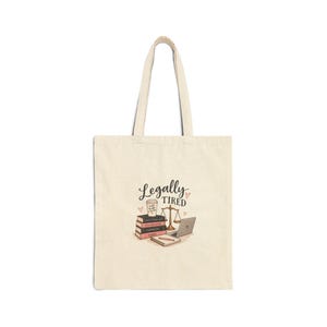 May include: A beige canvas tote bag featuring a graphic illustration of a stack of law books, a scale of justice, a laptop, a notebook with a pen, and a coffee cup. The text "Legally Tired" is printed in black script above the items.