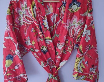 Op de afbeelding: Een rode badjas met bloemenprint en witte bies, hangend aan een houten hanger. De badjas heeft een levendig patroon van bloemen en bladeren in tinten blauw, geel en roze. De badjas is in de taille geknoopt.