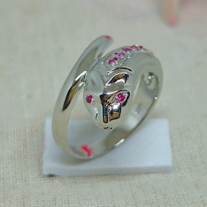 Könnte beinhalten: Ein silberner Ring mit einem Design aus rosafarbenen Steinen. Der Ring zeigt ein detailliertes Tierdesign mit rosafarbenen Steinen für die Augen. Der Ring befindet sich auf einem weißen Präsentationsständer.