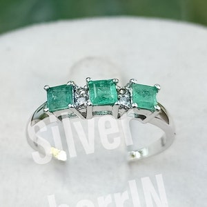 Puede incluir: Un anillo de plata con tres piedras preciosas cuadradas de color verde esmeralda y dos piedras preciosas más pequeñas transparentes.