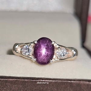 Peut inclure: Une bague en argent avec une grande pierre de rubis ovale entourée de deux petites pierres blanches rondes.