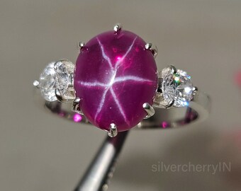 May include: A silver ring featuring a large, oval-shaped, pink star ruby gemstone and two smaller, round, white gemstones. The ring is set in a prong setting.