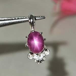 Puede incluir: Un colgante de plata con una piedra preciosa de rubí estrellado rosa con una inclusión en forma de estrella. El colgante está engastado con pequeñas piedras preciosas transparentes alrededor del rubí.