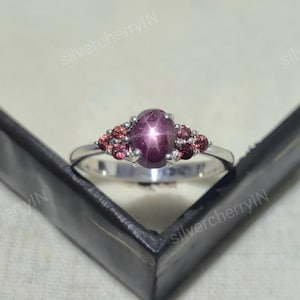 May include: A silver ring featuring a central oval cabochon-cut star ruby displaying a distinct six-rayed star effect. The center stone is flanked by three small round-cut reddish-pink gemstones on each side, all set in a polished silver band.