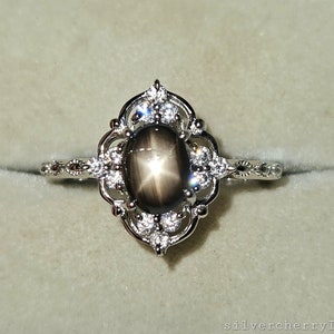 May include: A silver ring featuring a black star sapphire gemstone, encircled by smaller, clear gemstones. The ring has an elaborate, filigree design around the central stone and along the band. The ring is set against a light grey background.