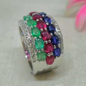 Può includere: Un anello d'argento con una fascia di pietre preziose smeraldo, rubino e zaffiro. L'anello è incastonato con piccoli diamanti attorno alle pietre preziose.