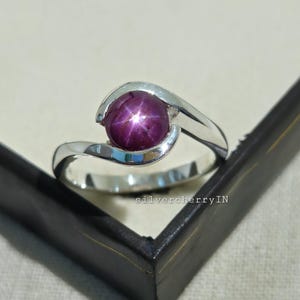 Peut inclure: Bague en argent avec une grosse pierre de rubis étoilé violet foncé. La bague a un design moderne avec une bande tourbillonnante qui maintient la pierre précieuse. La bague est présentée dans un écrin noir.