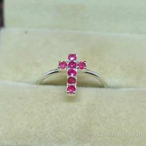 Op de afbeelding: Een zilveren ring met een rood robijn kruisontwerp. De ring bevindt zich in een kleine fluwelen doos.