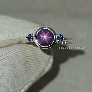 May include: A silver ring featuring a large, round, purplish-red star sapphire gemstone, flanked by two smaller blue gemstones. The ring has a simple, elegant design, perfect for everyday wear.