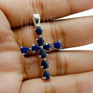 May include: A silver cross pendant with blue gemstone accents. The cross is adorned with teardrop-shaped stones, creating a delicate and elegant design.