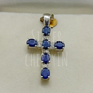 May include: A silver cross pendant with blue oval sapphires and smaller clear diamonds set in the metal. The pendant is in a white box.