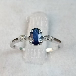 Puede incluir: Un anillo de plata con una piedra de zafiro ovalada azul y piedras más pequeñas transparentes en la banda. El anillo está en un expositor blanco. "SILVER CHERRYIN" está impreso en el expositor.
