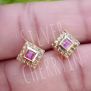 May include: A pair of gold square stud earrings with a pink square gemstone in the centre of each earring. The earrings are surrounded by a halo of smaller clear gemstones.
