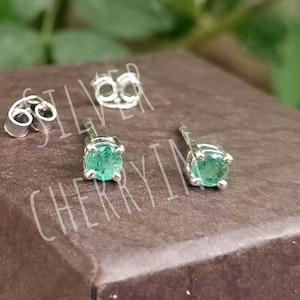 Könnte beinhalten: Ein Paar silberne Ohrringe mit grünen Edelsteinen. Die Ohrringe liegen auf einer braunen Oberfläche mit dem Text "SILVER CHERRY".