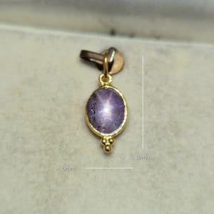 May include: A gold pendant featuring an oval-shaped purple star gemstone set in a gold bezel with a small decorative bead detail at the bottom. The pendant measures 8mm in width and 19mm in total height, including the top bail attachment.