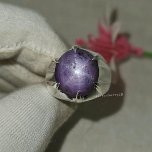 May include: A silver ring featuring a large, oval-shaped, purple gemstone with a star-like effect. The ring has a simple, modern design with claw-like prongs holding the stone. The background includes a soft, white fabric and a blurred pink flower.