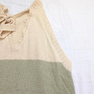 May include: A cream-coloured knitted top with a V-neck and a bow detail at the back. The top features horizontal stripes in shades of green and brown. The sleeveless design is visible in the image.