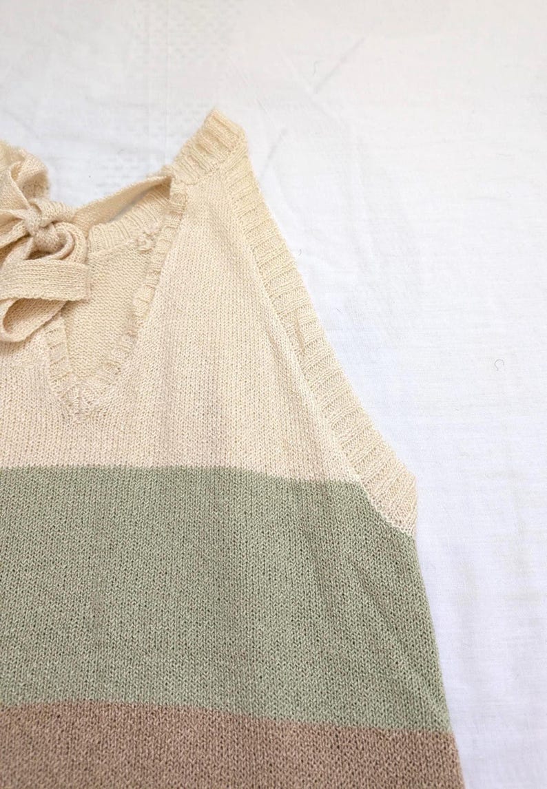 May include: A cream-coloured knitted top with a V-neck and a bow detail at the back. The top features horizontal stripes in shades of green and brown. The sleeveless design is visible in the image.