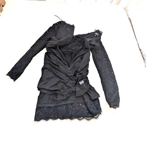 May include: Black dress with long lace sleeves and a layered skirt. The dress features a draped bodice with a zipper and a small label. The skirt has a lace trim. The dress is laid out on a white surface.