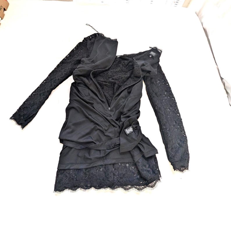 May include: Black dress with long lace sleeves and a layered skirt. The dress features a draped bodice with a zipper and a small label. The skirt has a lace trim. The dress is laid out on a white surface.