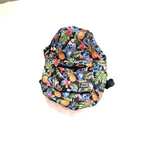 May include: Black backpack with a tropical print featuring pineapples, flowers, and the character Stitch. The backpack has a front pocket with a zipper and a black handle. The Disney Loungefly logo is visible on the front.