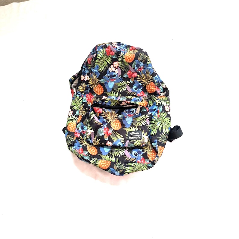 May include: Black backpack with a tropical print featuring pineapples, flowers, and the character Stitch. The backpack has a front pocket with a zipper and a black handle. The Disney Loungefly logo is visible on the front.