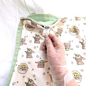Op de afbeelding: Een witte babydeken met een herhaald patroon van het personage Grogu uit de Star Wars-serie, met een lichtgroene rand. De deken heeft een label met het Star Wars-logo.