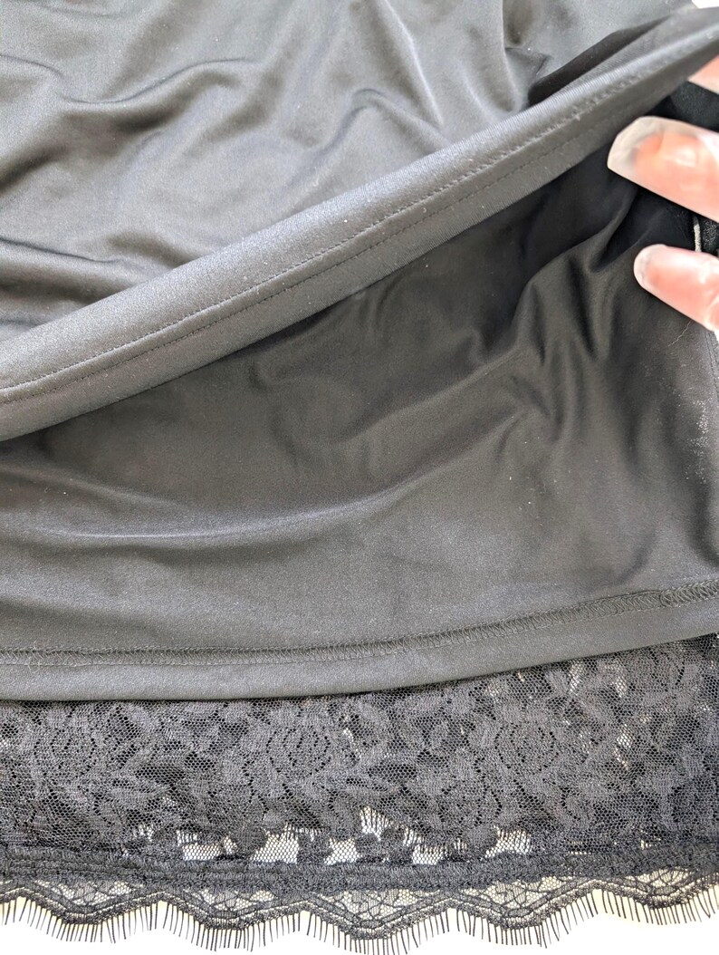 May include: Black slip dress with a lace hem. The dress features a smooth, shiny fabric and a delicate lace trim at the bottom. The image shows a close-up of the dress's details, highlighting the texture and design.