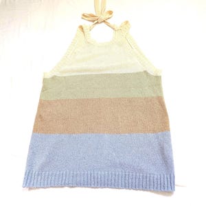 May include: A knitted halter top with a striped pattern. The top has a cream-coloured upper section, with stripes of green, tan, and light blue below. The halter straps are tied in a bow at the back of the neck. The garment is made of a knitted material.