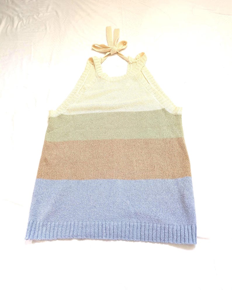 May include: A knitted halter top with a striped pattern. The top has a cream-coloured upper section, with stripes of green, tan, and light blue below. The halter straps are tied in a bow at the back of the neck. The garment is made of a knitted material.