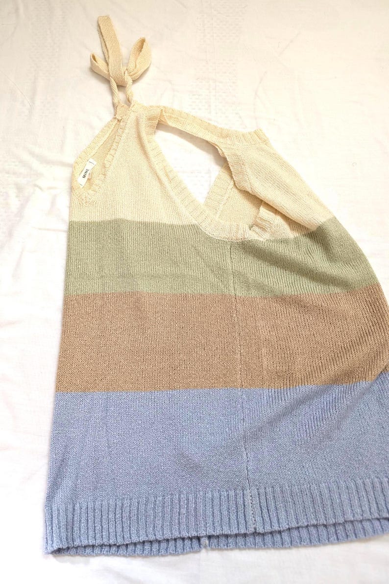 May include: A knitted top in a striped pattern of cream, olive green, tan, and light blue. The top has a halter neck design with tie straps and a V-neckline. The fabric appears soft and lightweight, suitable for warmer weather.
