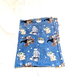 May include: A folded blue blanket featuring Star Wars characters. The blanket has a repeating pattern of Grogu, R2-D2, and the Mandalorian, along with white stars. The blanket is made of a soft, plush material.