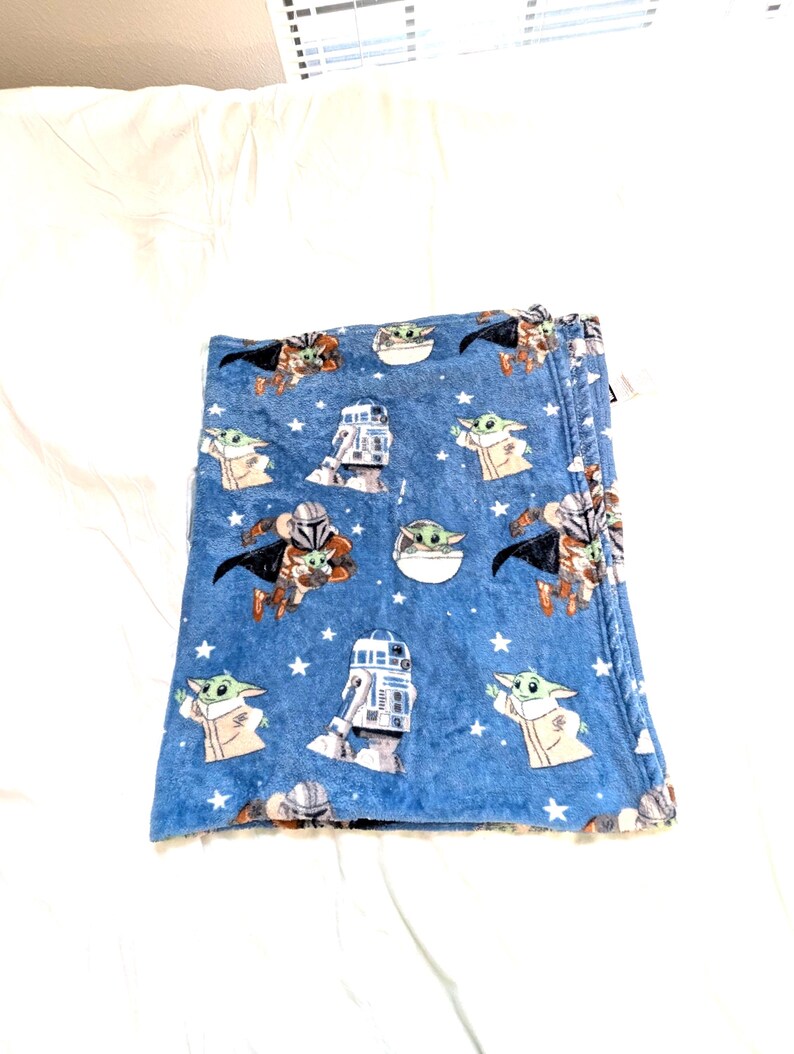 May include: A folded blue blanket featuring Star Wars characters. The blanket has a repeating pattern of Grogu, R2-D2, and the Mandalorian, along with white stars. The blanket is made of a soft, plush material.