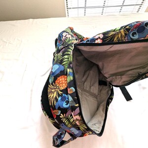 May include: Black backpack with a colorful tropical print featuring Stitch from Lilo & Stitch, pineapples, and flowers. The backpack has a large main compartment and a smaller front pocket. The interior is lined with a striped fabric.