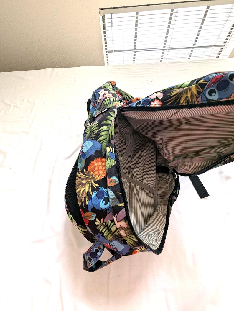 May include: Black backpack with a colorful tropical print featuring Stitch from Lilo & Stitch, pineapples, and flowers. The backpack has a large main compartment and a smaller front pocket. The interior is lined with a striped fabric.
