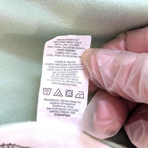 Op de afbeelding: Een close-up van een kledinglabel met wasvoorschriften in het Engels en Spaans. Het label bevat symbolen voor wassen, drogen en strijken. De fabrikant is Bentex Group, Inc., geproduceerd in Zaozhuang, China, in oktober 2020.