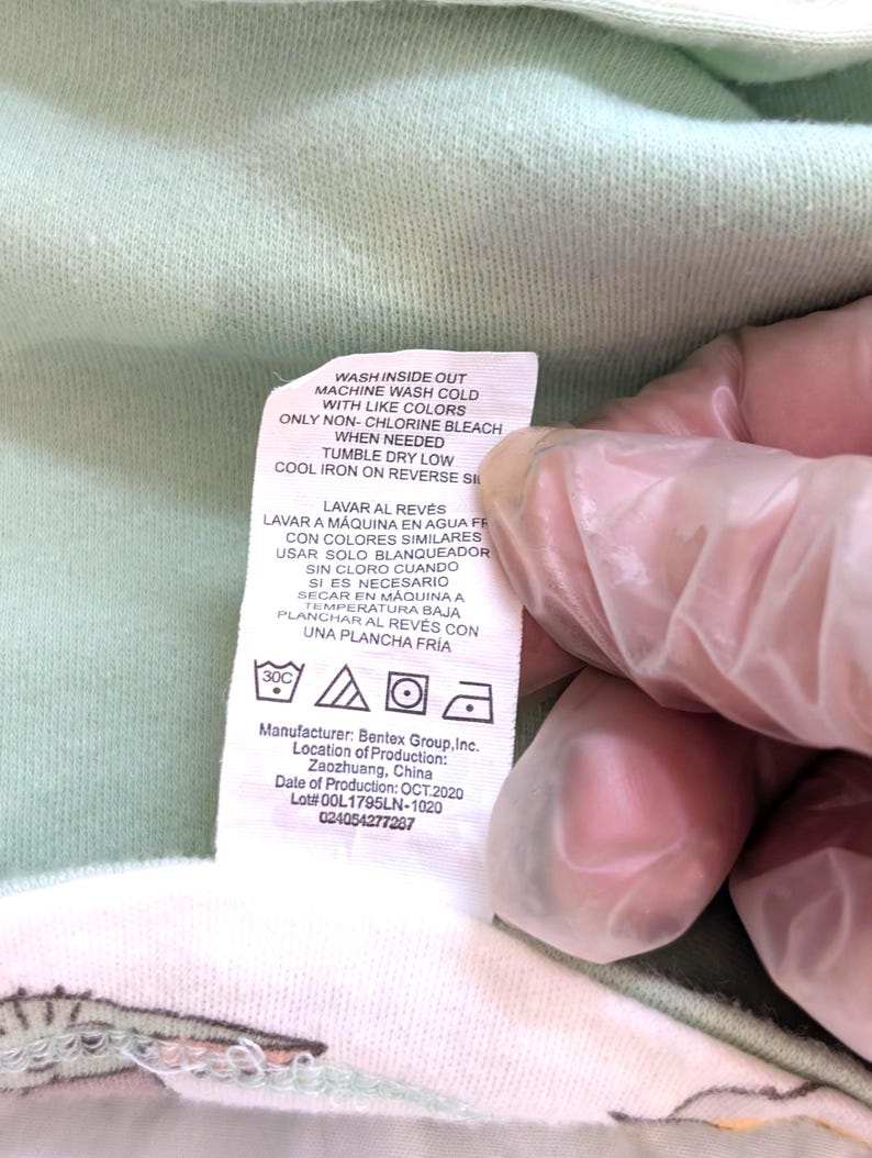 Op de afbeelding: Een close-up van een kledinglabel met wasvoorschriften in het Engels en Spaans. Het label bevat symbolen voor wassen, drogen en strijken. De fabrikant is Bentex Group, Inc., geproduceerd in Zaozhuang, China, in oktober 2020.