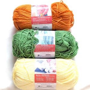 May include: Three skeins of yarn in orange, green, and yellow. Each skein has a label with text and a picture of a knitted item. The yarn is from the brand Bernat. The yarn is for knitting and crochet projects.