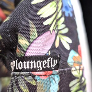May include: Close-up of a black fabric with a colorful floral pattern. The design includes green leaves, pink flowers, and blue accents. A black label with the white text "Loungefly" is sewn onto the fabric.