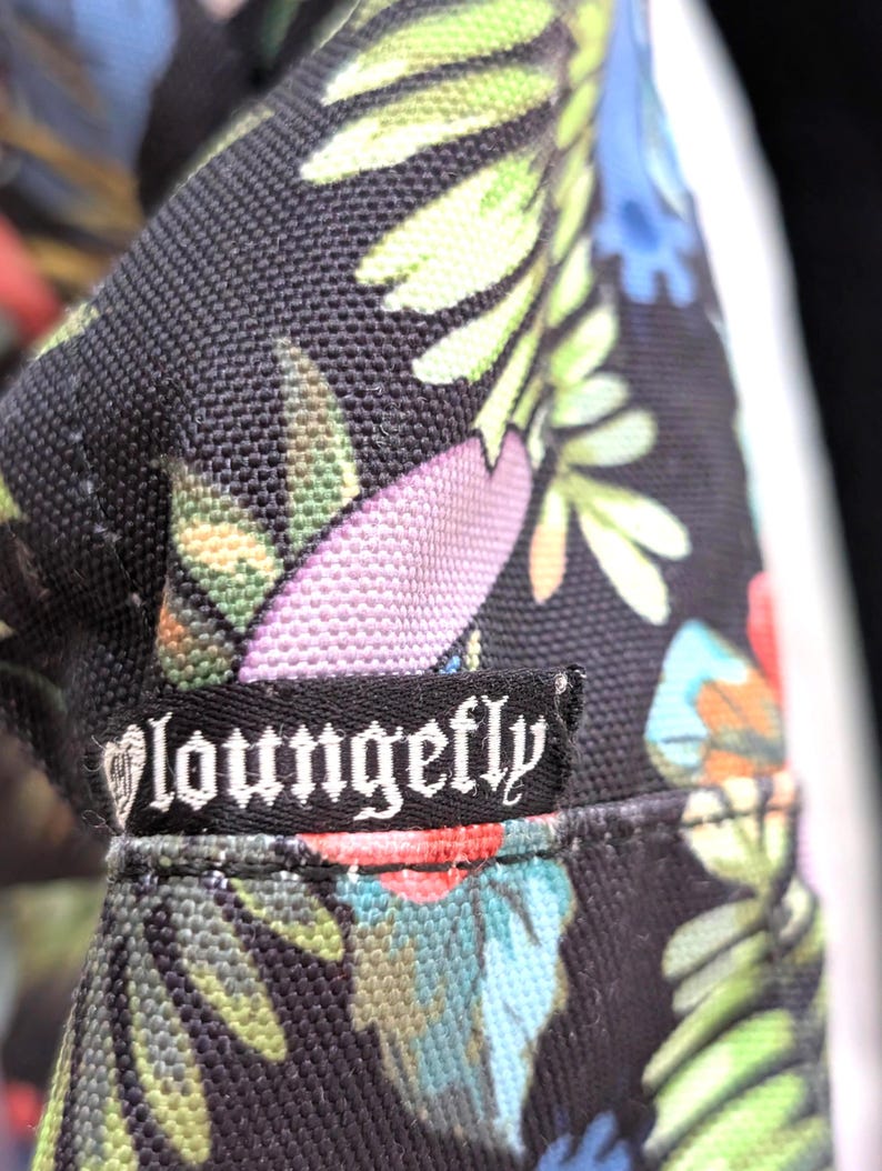 May include: Close-up of a black fabric with a colorful floral pattern. The design includes green leaves, pink flowers, and blue accents. A black label with the white text "Loungefly" is sewn onto the fabric.