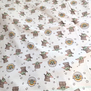 Op de afbeelding: Wit T-shirt met een herhaald patroon van Grogu, ook bekend als Baby Yoda, uit de Star Wars-serie. Het ontwerp bevat Grogu in verschillende poses, samen met sterren. Het shirt heeft een lichtgroene rand.