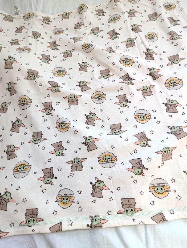 Op de afbeelding: Wit T-shirt met een herhaald patroon van Grogu, ook bekend als Baby Yoda, uit de Star Wars-serie. Het ontwerp bevat Grogu in verschillende poses, samen met sterren. Het shirt heeft een lichtgroene rand.