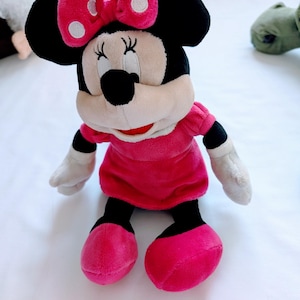 May include: A plush Minnie Mouse doll with a pink dress and bow. The doll has black ears, a black nose, and white gloves. The bow features white polka dots. The doll's feet are a vibrant pink, and the eyes are closed.